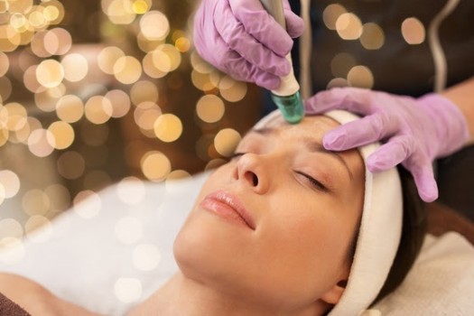 Woman laying with her head on a table while a girl with purple gloves gives her a chemical peel.